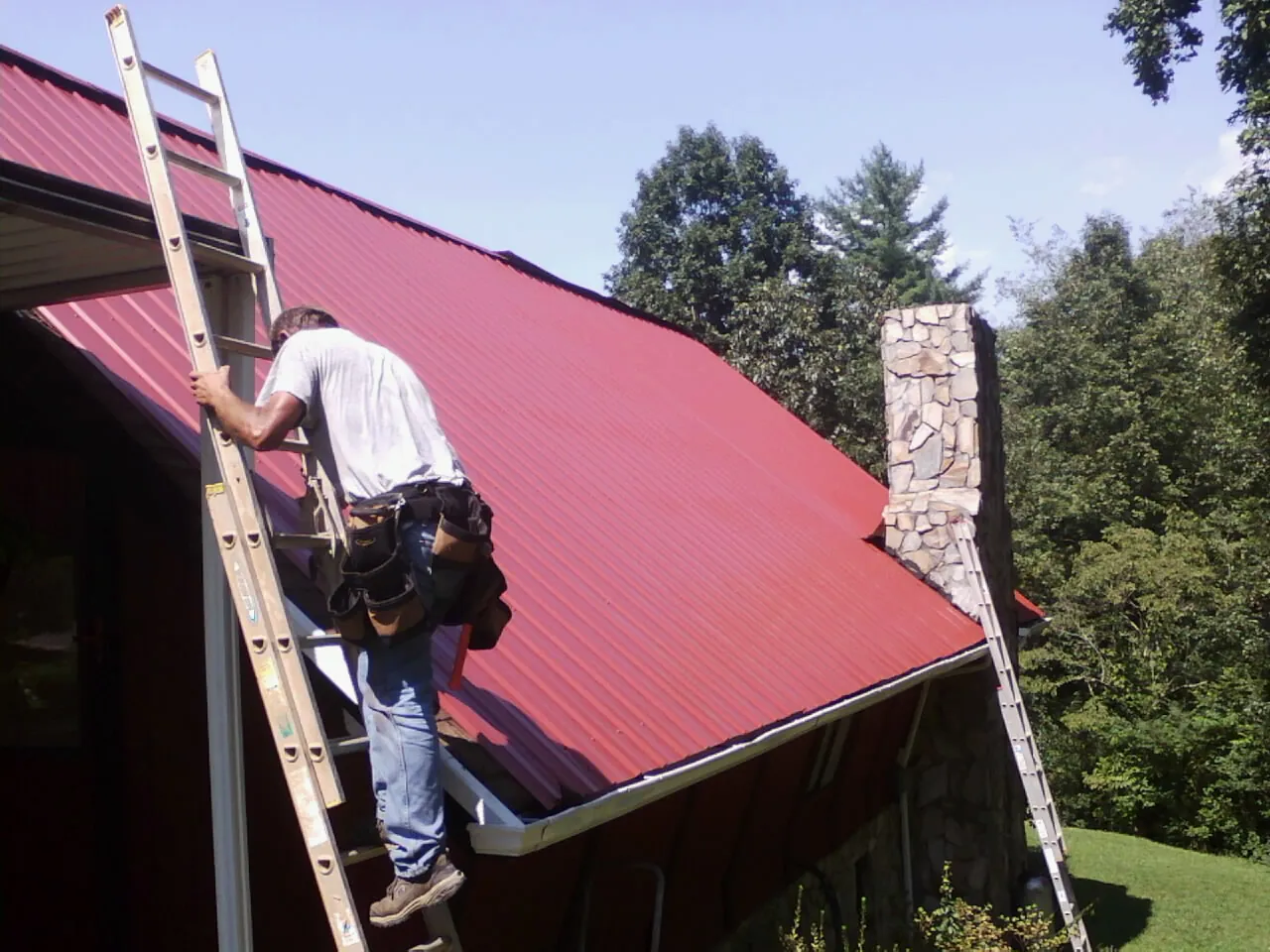 Roofer on a ladder beside a red metal roof and stone chimney — Soffit & Fascia Repair by Roofing Brothers in San Gabriel